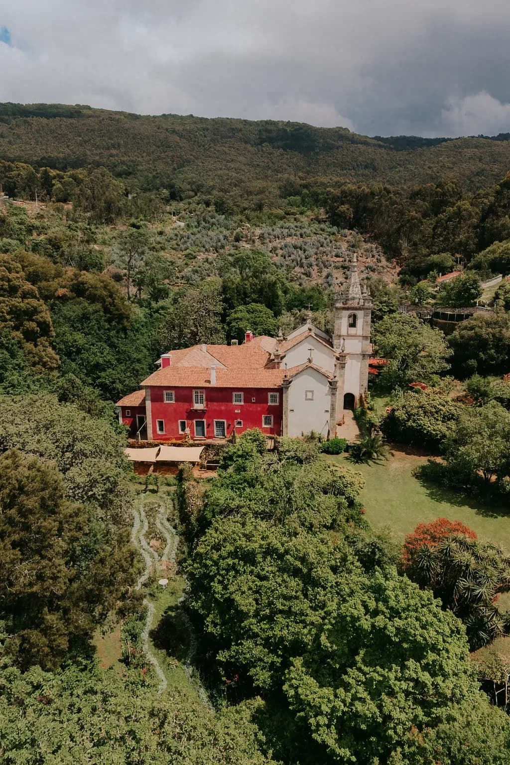 La maison rouge et l’église vues du ciel, la montagne boisée derrière et les allées du jardin en contrebas