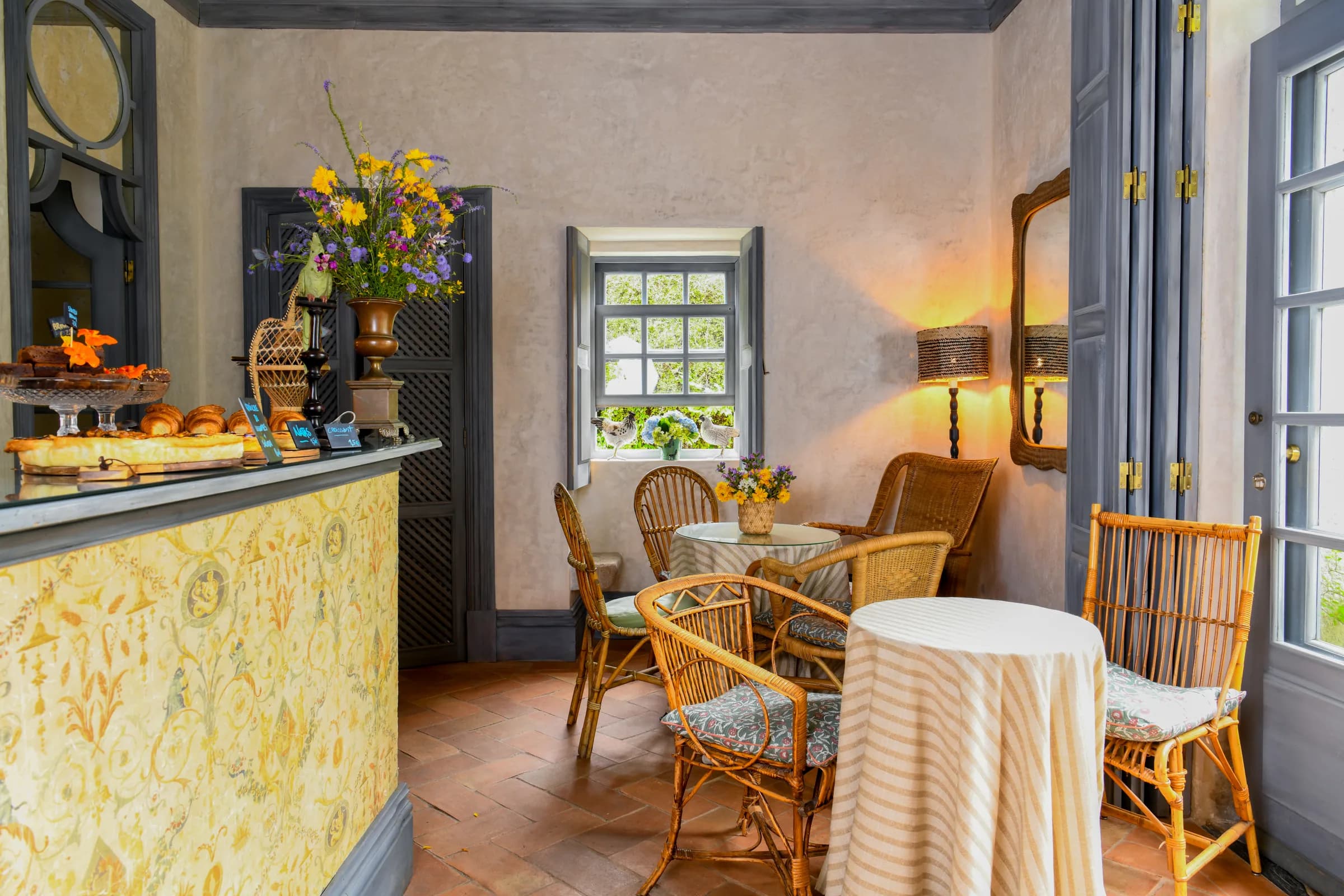 The café counter dressed with wildflowers and pastries, wicker chairs by the open window