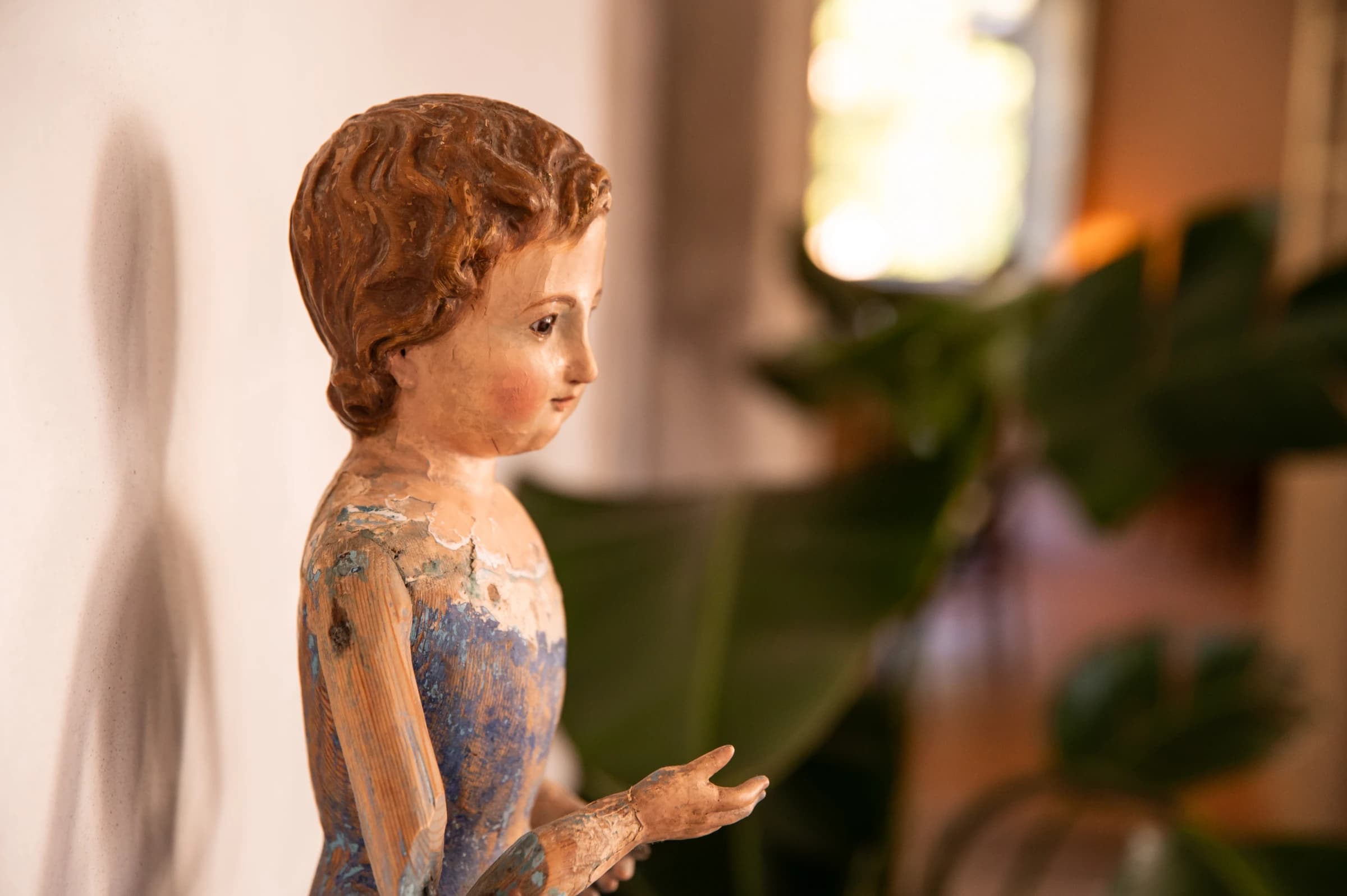 A weathered polychrome wood statue of a child, greenery softly out of focus behind it