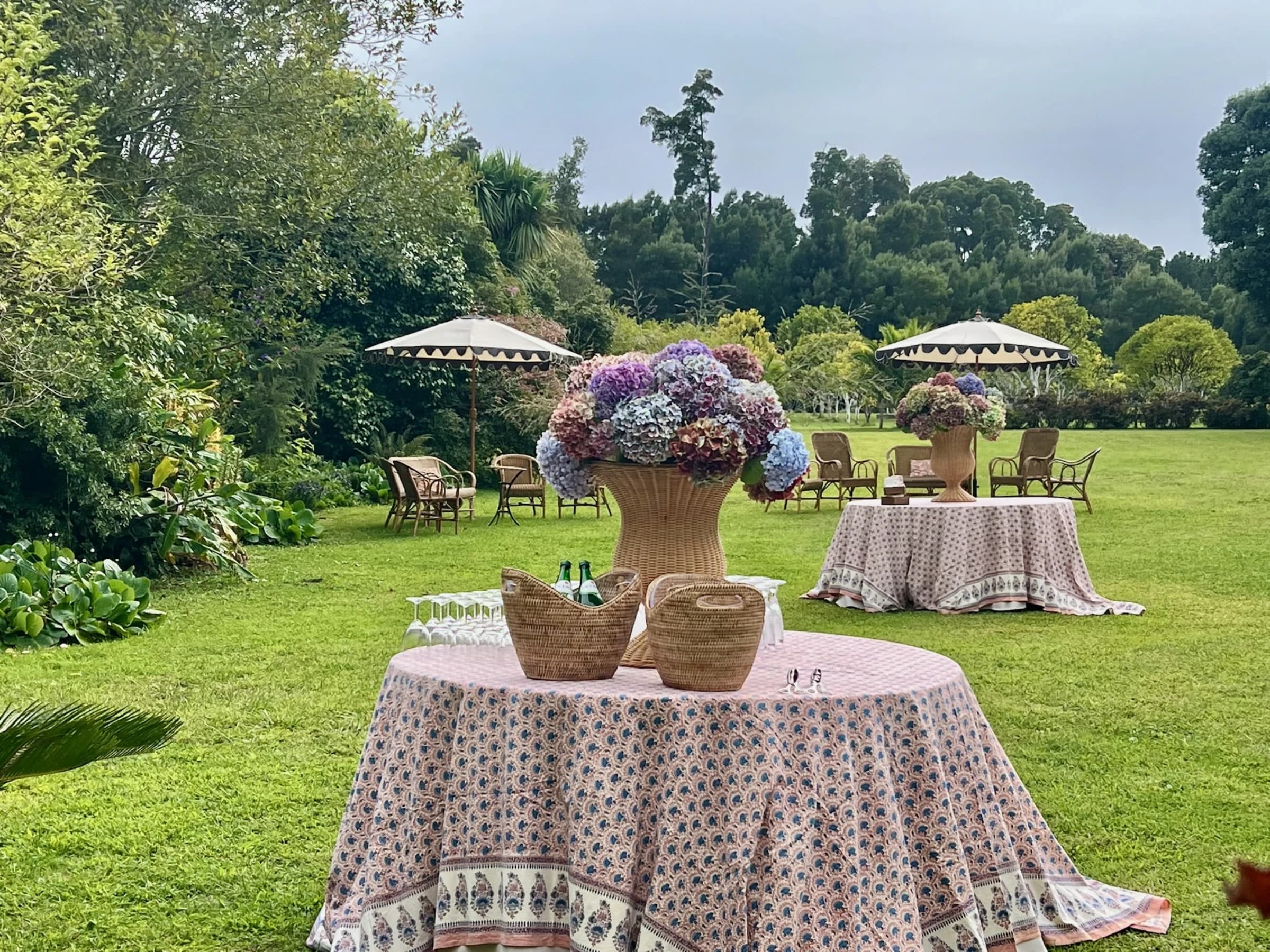 Tables dressed in floral linen beneath garden umbrellas, hydrangeas in woven urns, on the estate's open lawn