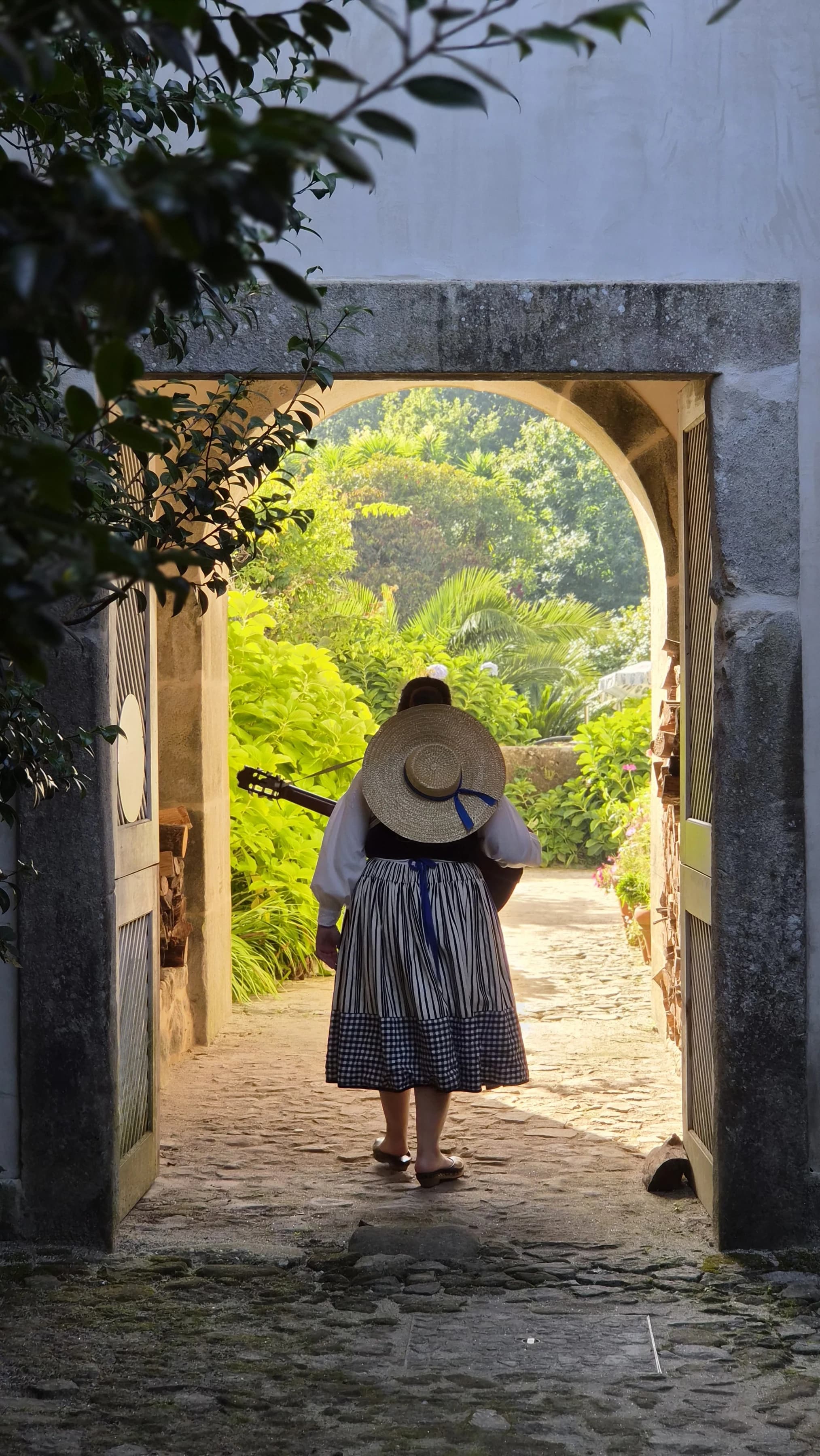 A folk musician in traditional Minho dress, guitar in hand, crossing a garden archway