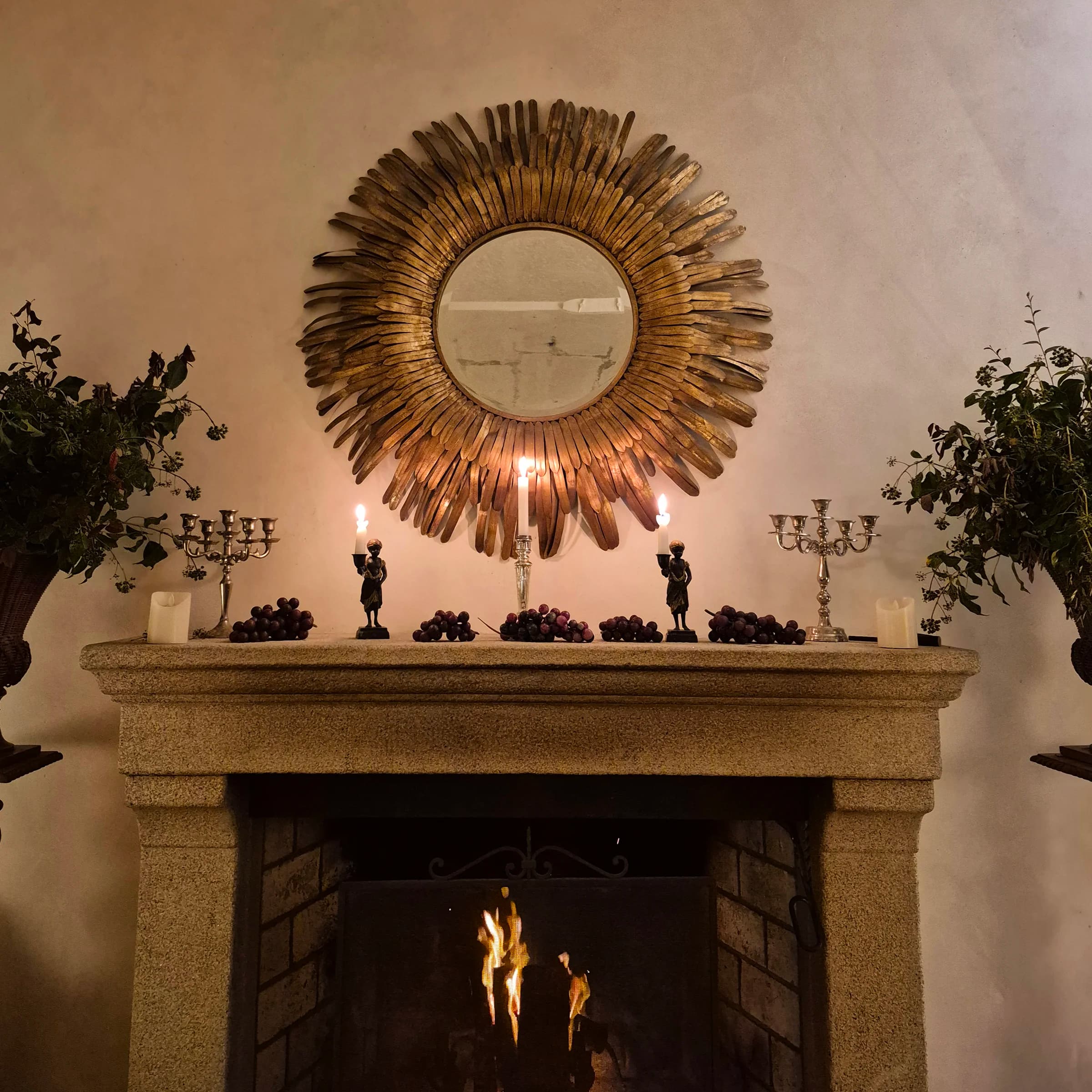 The stone mantelpiece dressed with candles and grapes beneath a gilded sunburst mirror